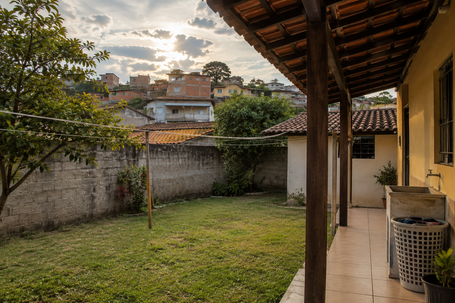 Quintal residencial com varal vazio e cesto sob uma varanda coberta, com casas ao fundo e céu entre nuvens.